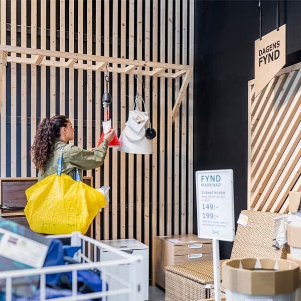 Žena u IKEA robnoj kući razgledava lampe, drži žutu vrećicu, pored nje je znak s cijenama.