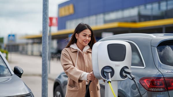 Žena v béžovém kabátu před obchodním domem, která používá nabíjecí stanici pro své auto.