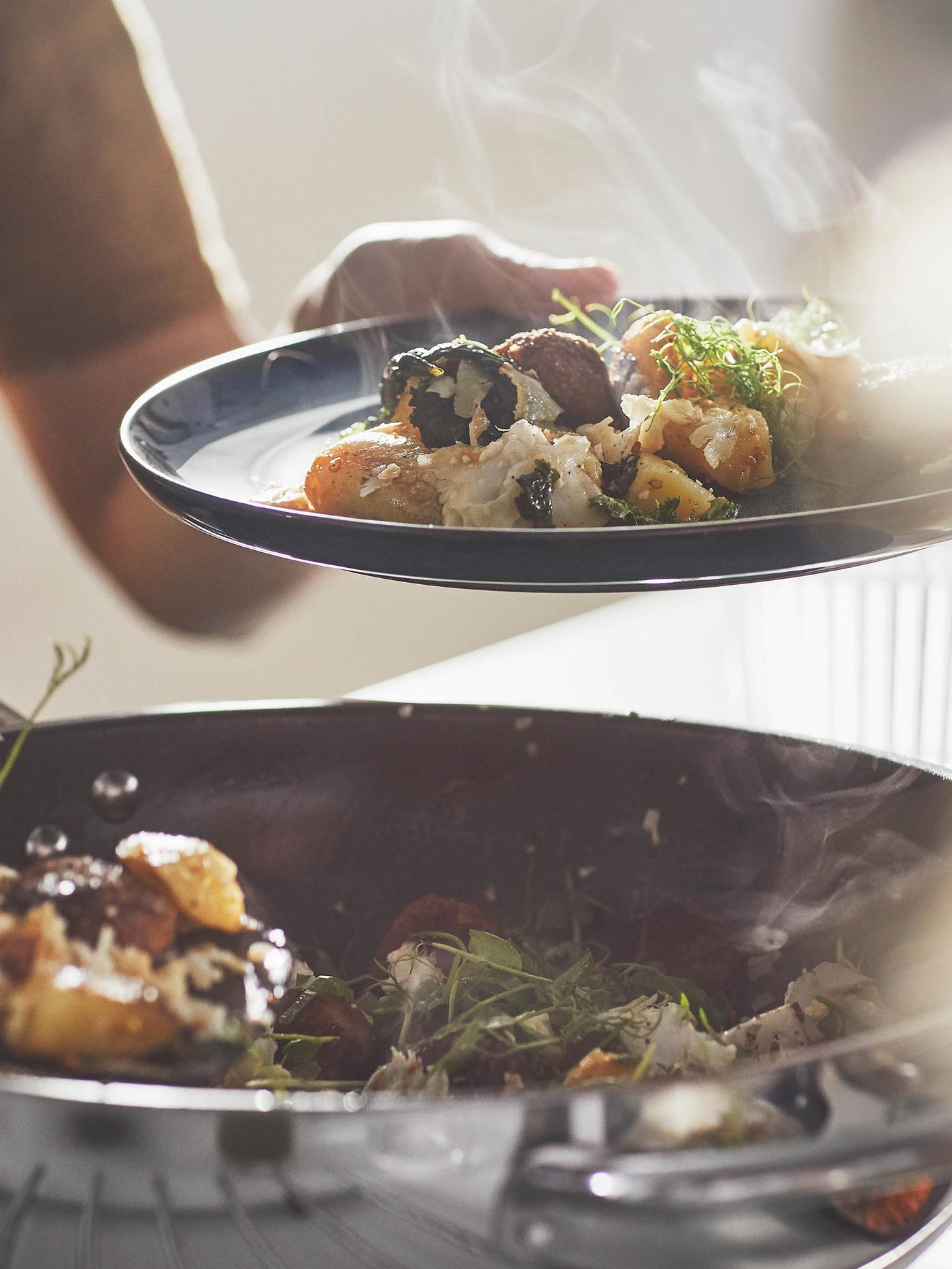 Žena v béžovém svetru drží talíř se směsí kuliček a zeleniny nad pánví wok IKEA 365+.