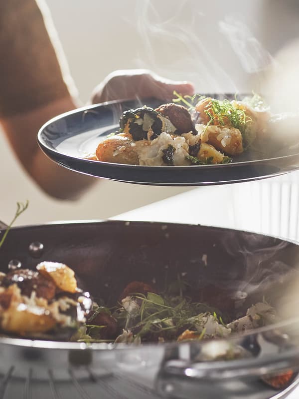 Žena v béžovém svetru drží talíř se směsí kuliček a zeleniny nad pánví wok IKEA 365+.