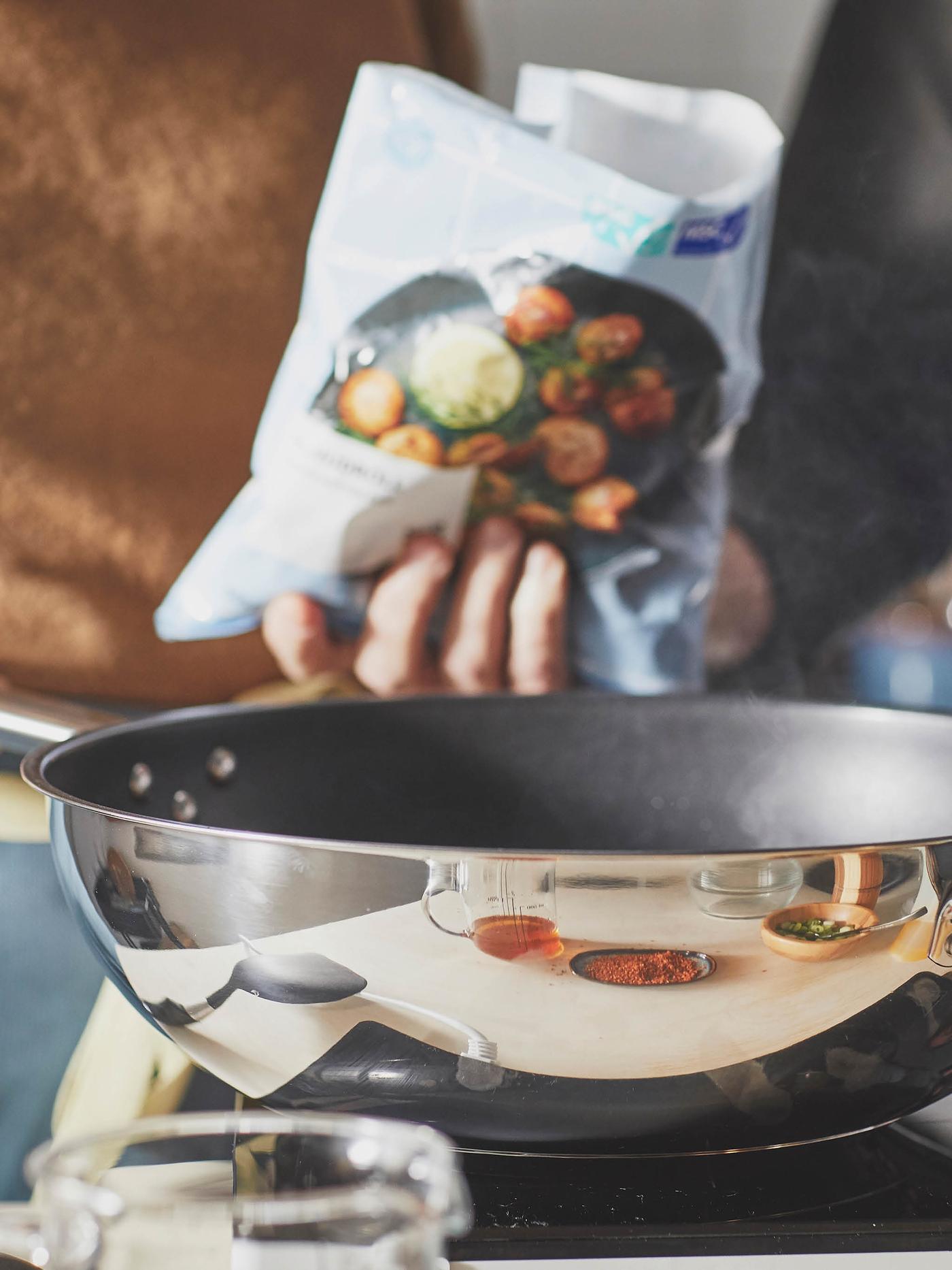 Žena v béžovom svetri drží vrecko rybacích guľôčok HUVUDROLL pred panvicou wok IKEA 365+.