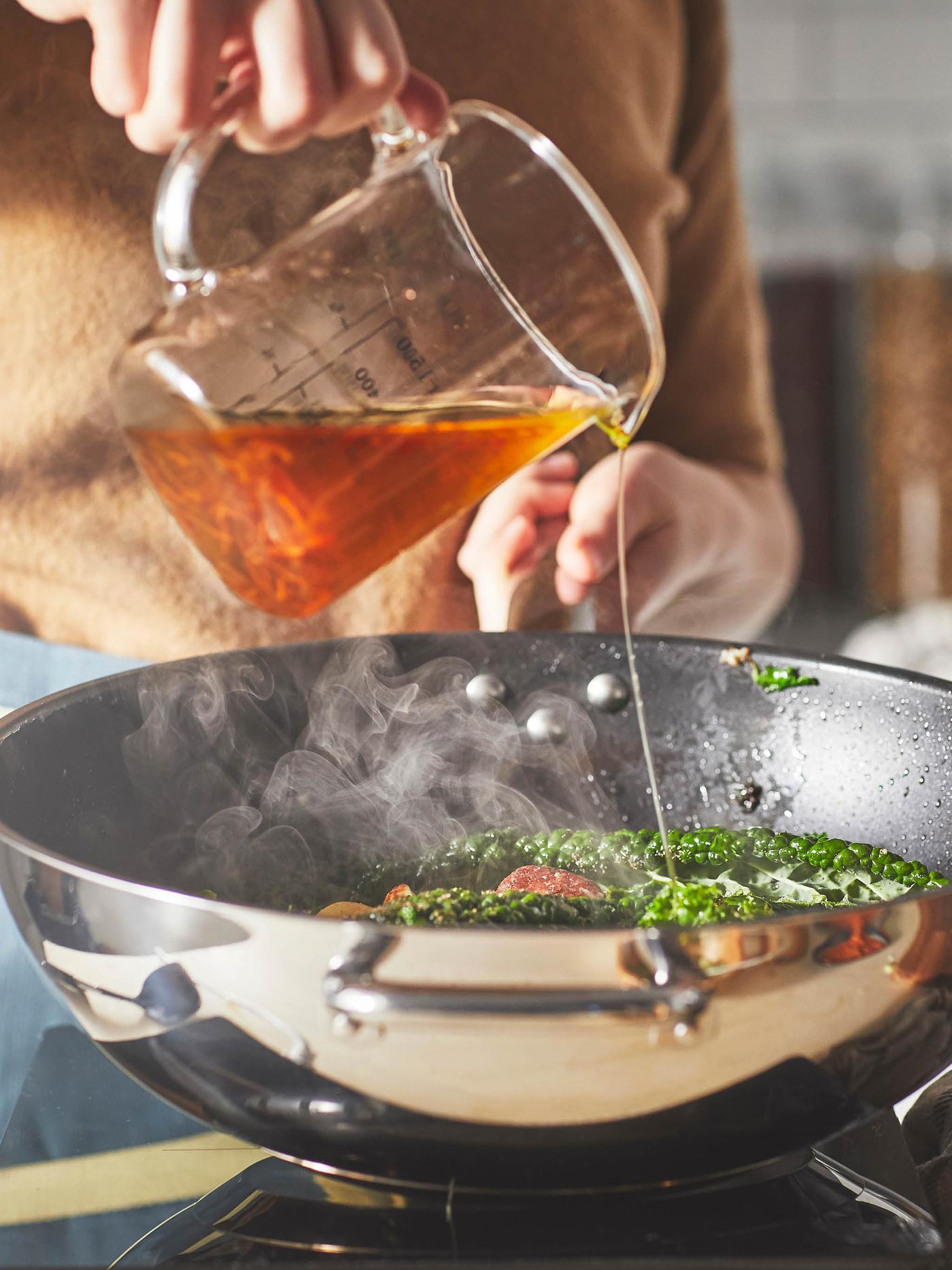 Žena v béžovom svetri leje vývar z odmerky VARDAGEN do panvice wok IKEA 365+ na zmes zeleniny.