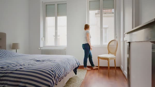 Žena v džínách a tričku stojí a dívá se z okna své ložnice.