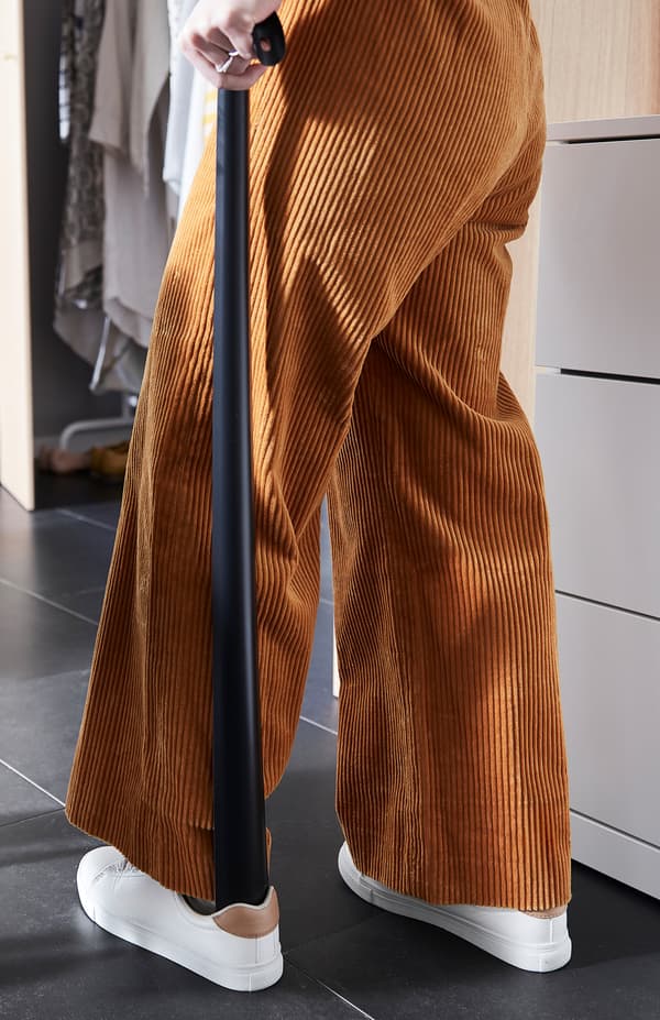 Žena v menčestrových nohaviciach používa obuvák OMTÄNKSAM na obúvanie po stojačky.