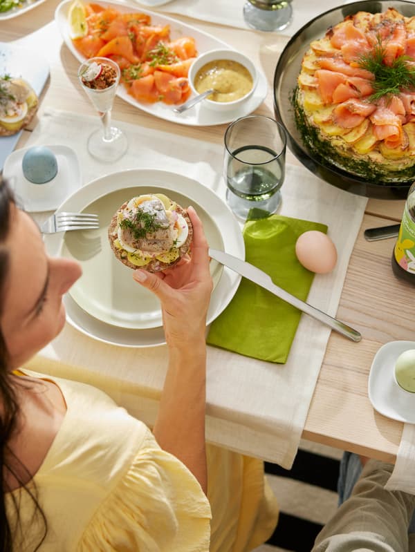 Žena ve žlutých šatech drží v ruce knäckebrot s vajíčky a naloženými sledi, před ní na stole jsou zapečené brambory s lososem a uzený losos s hořčičnou omáčkou.