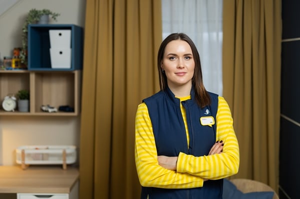 Женщина в форме IKEA стоит, скрестив руки, в интерьере с полками и шторами на заднем плане.