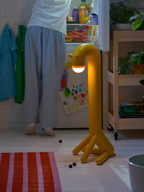 Žltá stojacia lampa v tvare žirafy GREJSIMOJS stojí v kuchyni, za ňou niekto pátra v otvorenej chladničke.
