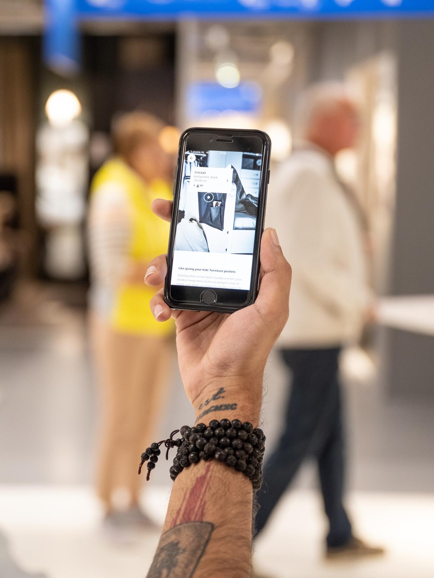 Žmogus stovi parduotuvėje ir rankoje laiko išmanųjį telefoną su įjungta IKEA programėle; fone matosi kiti pirkėjai.