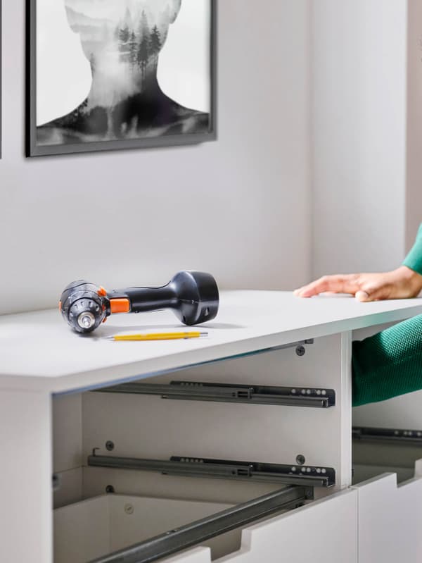 Zöld felsőt viselő ember, amint egy FIXA fúrót használva éppen a falhoz rögzíti a fehér NORDLI szekrényt.