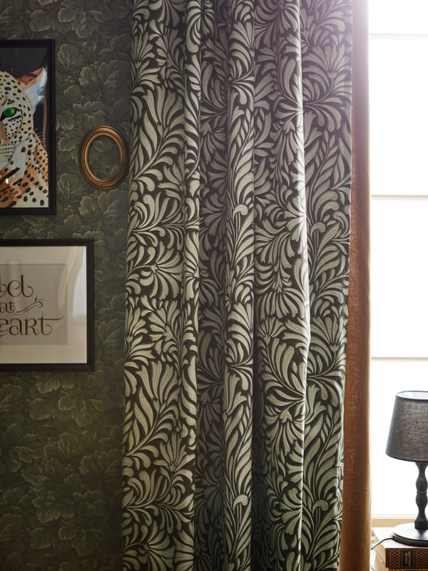 Zöld mintás TRÅGSPINNARE függönyt látunk egy természetes zöld mintás tapétás fal előtt.