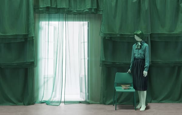 Zöld ruhába öltöztetett figura áll egy zöld szék mellett, zöld függönyök előtt.