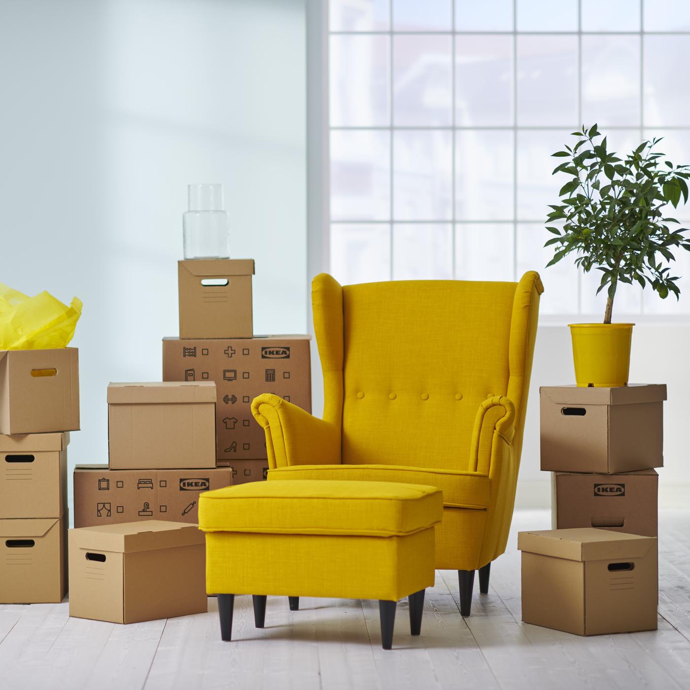 Żółtry fotel STRANDMON z podnóżkiem na tle kartonów ustawionych w stosy. 