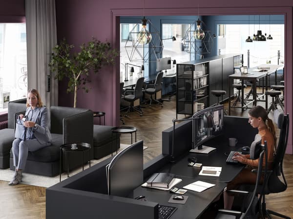 Zona de recepción de color ciruela con una mujer sentada en un módulo de asiento VALLENTUNA negro y otra mujer detrás del mostrador de recepción negro.