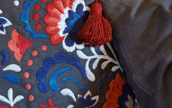 Zoomed-in section of a cushion cover, embroidered with intricate, different-colour patterns, a tassel hanging from a corner.