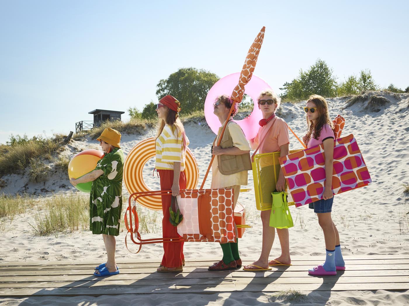 zu sehen 5 Personen am Strand, stehen hintereinander in einer Reihe: jede:r trägt ein Strandaccessoire, Wasserball, Liegestuhl, Strandsessel, Sonnenschirm - alle aus der neuen IKEA STRANDÖN Serie in orange, magenta und weiß. Fröhlich, bunt. 