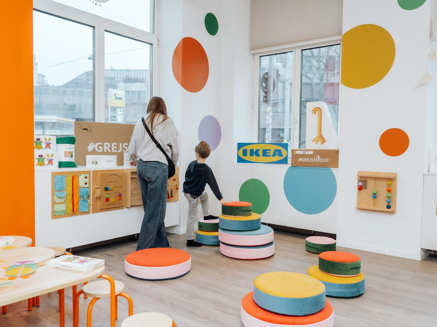 Zu sehen ein bunt gestalteter Raum mit zwei Fenstern, zwei Menschen in der Rückansicht, Mutter und Sohn, balancieren auf bunten IKEA Stapelsteinen der neuen GREJSIMOJS Kollektion. 