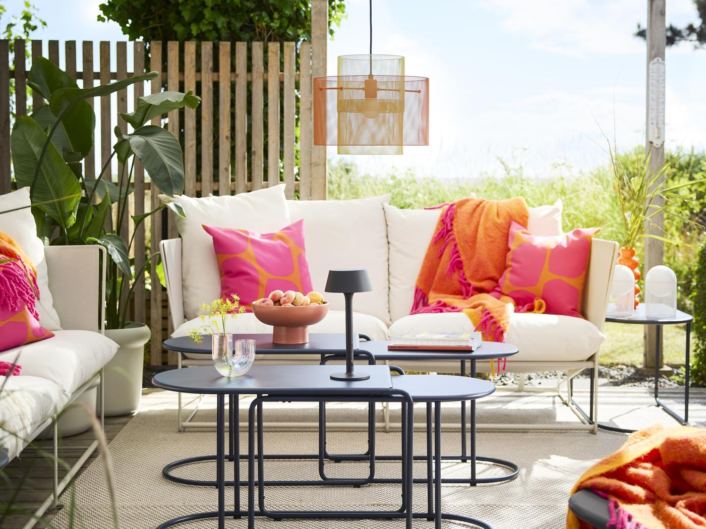 zu sehen eine Terrasse mit weißen Sofaelementen, bunten (pink orangen) Outdoorkissen und schwarzen runden Satztischen. 