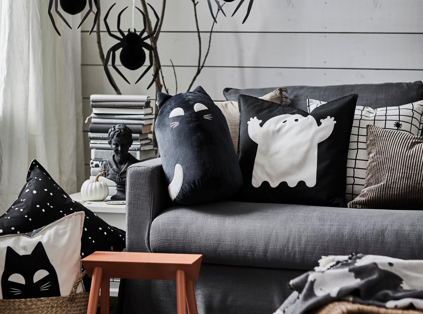 Zu sehen ist ein Ausschnitt eines Wohnzimmers: eine grau Couch, darauf verschiedene Kissen mit Halloween-Motiven. Im Hintergrund Hängedeko-Artikel in Form von Spinnen. 