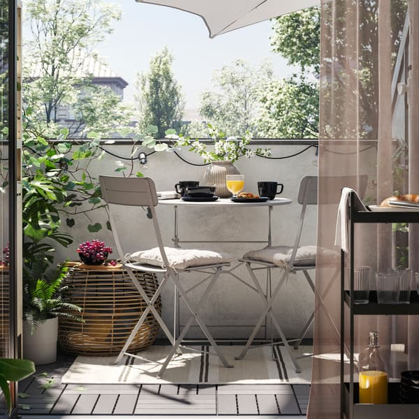 Zu sehen ist ein Balkon mit einem grauen SUNDSÖ Tisch für draußen und zwei grauen Stühlen auf einem Teppich, u. a. mit einem Couchtisch, Pflanzen und einem Sonnenschirm.