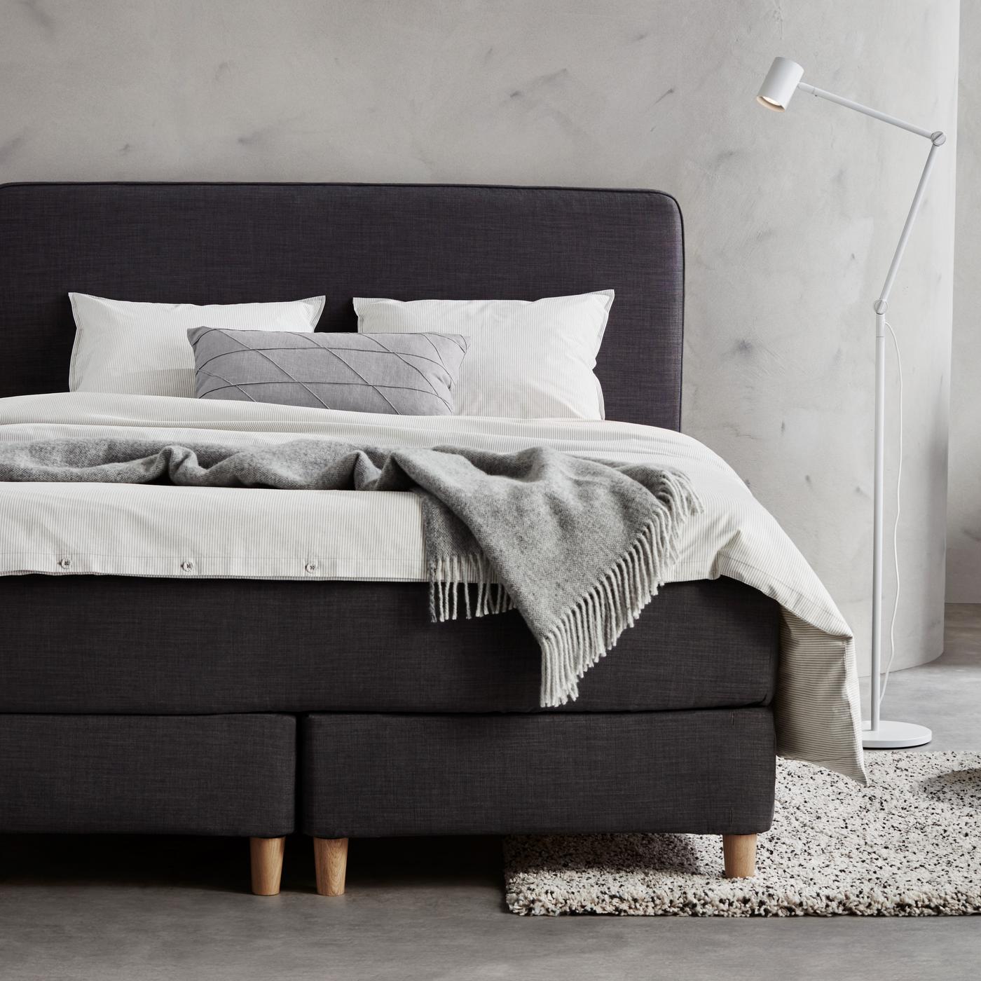 Zu sehen ist ein dunkelgraues Boxspringbett mit weißer Bettwäsche und einem hellgrauen Plaid.