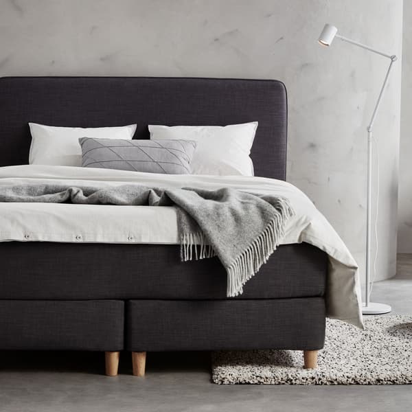 Zu sehen ist ein dunkelgraues Boxspringbett mit weißer Bettwäsche und einem hellgrauen Plaid.
