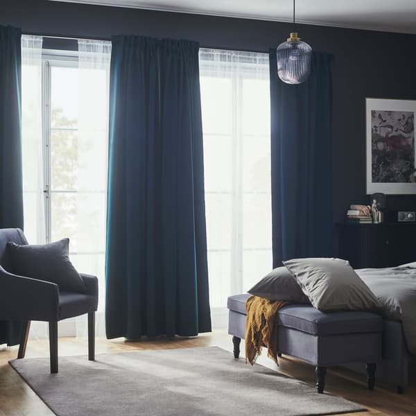 Zu sehen ist ein Schlafzimmer mit MAJGULL verdunkelnden Gardinen in Dunkeltürkis und Netzgardinen an grossen Fenstern. In der Nähe stehen ein Bett und eine Bank.