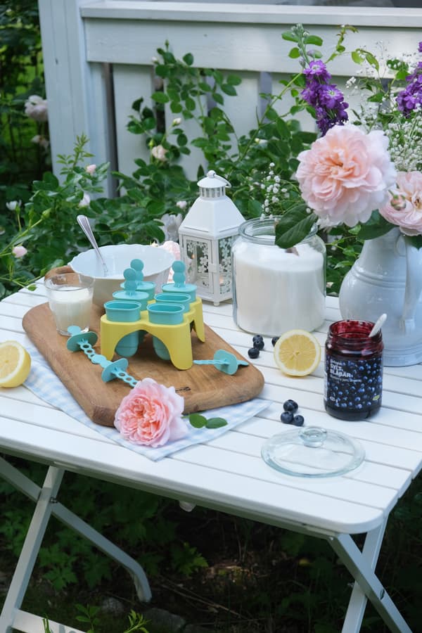 Zu sehen ist ein weißer Tisch in einem Garten, auf dem verschiedene Getränke und Speisen für den Sommer zu sehen sind.
