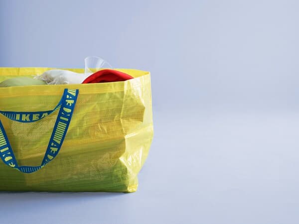 Žuta IKEA torba s plavim natpisom, napunjena crvenim i bijelim predmetima, na svijetloplavoj pozadini.