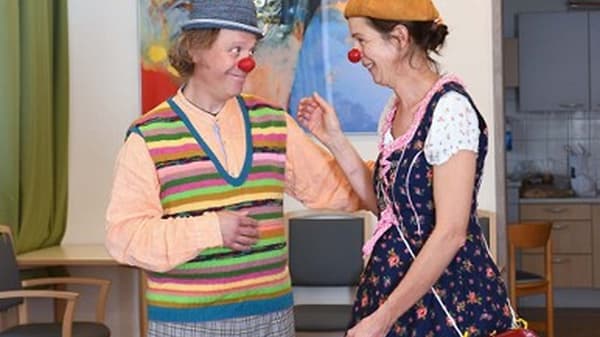 Zwei als Klinikclowns verkleidete Personen stehen in einem Gemeinschaftsraum. Beide tragen rote Clownnasen und farbenfrohe Kleidung. Eine Person trägt eine gestreifte Weste und eine Kappe, die andere ein gemustertes Kleid mit Mütze. Im Hintergrund sind Stühle, ein Tisch und ein Bild an der Wand zu sehen.