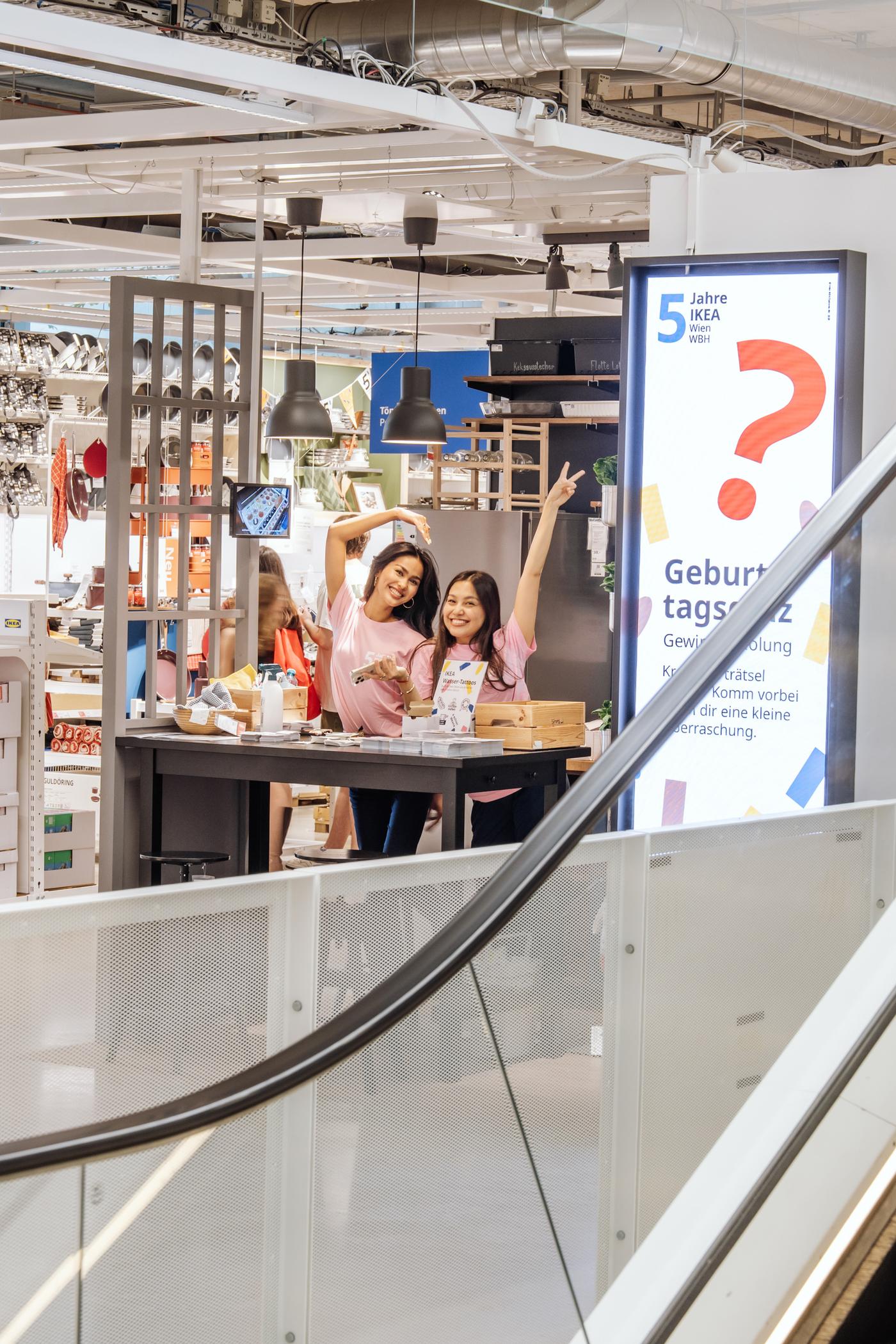Zwei Besucherinnen winken von einer Rolltreppe im IKEA Einrichtungshaus.