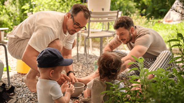 Zwei Erwachsene knien im Garten auf Kiesboden und spielen mit zwei Kleinkindern bei sommerlichem Wetter. Im Hintergrund stehen Gartenmöbel und viel Grün.