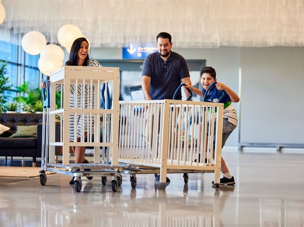 Zwei Erwachsene und ein Kind, die ein Gitterbett und eine Wickelkommode auf einem Wagen in ein IKEA Einrichtungshaus schieben.