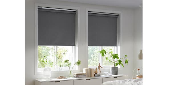 Zwei Fenster mit heruntergelassenen grauen Rollos in einem hellen Schlafzimmer mit weißen Möbeln und grünen Zimmerpflanzen auf der Kommode.