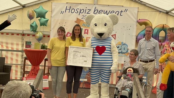 Zwei Frauen in gelben IKEA-Shirts und ein grosses Maskottchen in Bärenkostüm mit blau-weiss gestreiftem Anzug und rotem Herz auf der Brust stehen auf einer Bühne und halten einen Spendenscheck über 4'000 Euro von IKEA Wetzlar. Im Hintergrund ist ein Banner mit dem Schriftzug „Hospiz bewegt“ sowie bunte Ballons zu sehen. Eine Frau im Rollstuhl hält eine Stoffpuppe, während weitere Personen an der Veranstaltung teilnehmen.