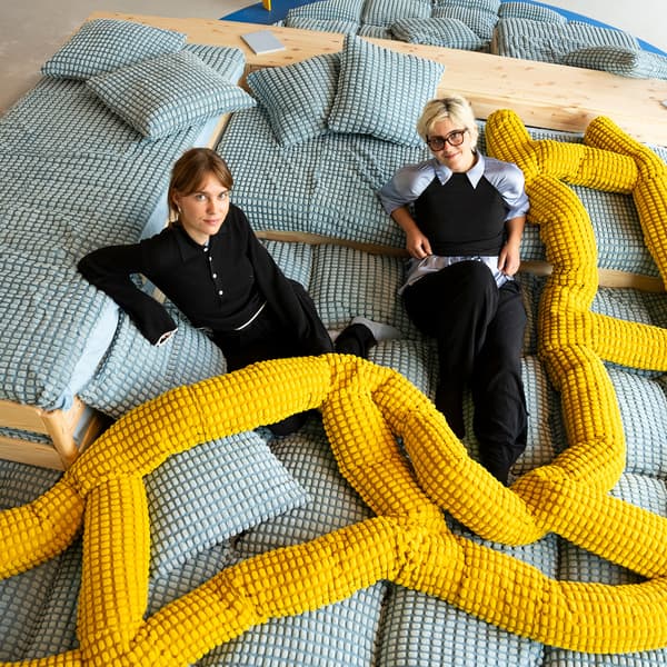 Zwei Frauen mit dunkler Kleidung die auf IKEA Pölstern sitzen. darüber eine leichte 
