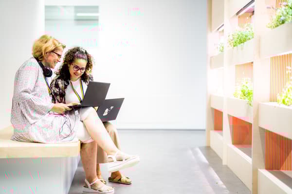Zwei Frauen sitzen auf einer Bank und arbeiten gemeinsam an einem Laptop in einem hellen, modernen Arbeitsbereich mit grünen Pflanzen im Hintergrund.