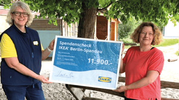 Zwei Frauen stehen draussen unter einem Baum und halten gemeinsam einen grossen Spendenscheck mit der Aufschrift „IKEA Berlin-Spandau“ über 11'900 Euro. Die Frau links trägt eine gelbe IKEA-Mitarbeiterkleidung mit blauer Weste und lächelt in die Kamera.