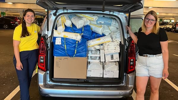 Zwei Frauen stehen in einem Parkhaus neben einem geöffneten Kofferraum, der randvoll mit IKEA-Spenden wie Kissen, Bettwaren, Tellern und blauen IKEA-Taschen gefüllt ist. Eine der Frauen trägt ein gelb gestreiftes T-Shirt, das auf eine IKEA-Mitarbeiterin hinweist. Beide lächeln in die Kamera.  