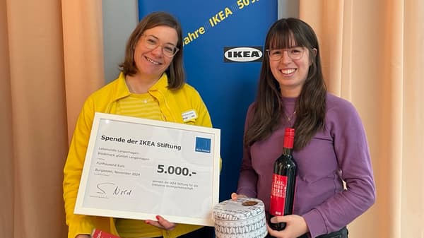 Zwei Frauen stehen vor einem blauen IKEA-Banner. Eine Frau hält einen Spendenscheck über 5.000 Euro, die andere hält eine Flasche Wein und eine kleine Dose mit Muster.