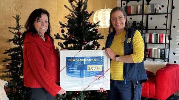 Zwei Frauen stehen vor einem weihnachtlich geschmückten Hintergrund mit Weihnachtsbäumen und Lichterketten. Sie halten gemeinsam einen IKEA-Spendenscheck über 3.000 Euro.