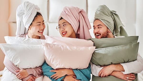 Zwei Frauen und ein Mann sitzen lächelnd, halten alle je zwei Kissen in der Hand und haben ein GULVIAL Handtuch farblich passend zu den Kissen als Turban auf dem Kopf.