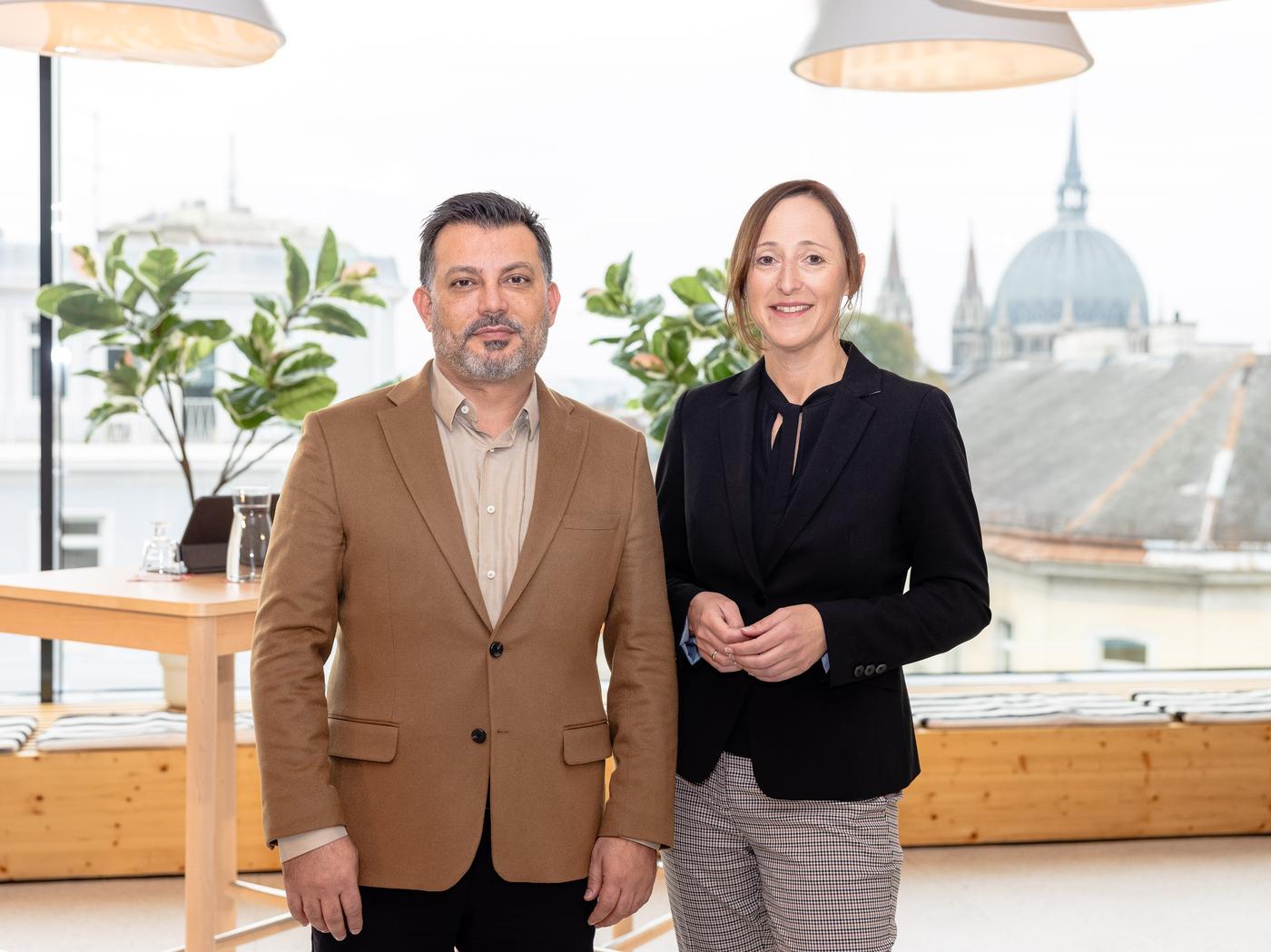 Zwei Führungskräfte von IKEA stehen in einem modernen Besprechungsraum mit Blick über die Dächer einer Stadt – professionelles Team im urbanen Arbeitsumfeld.

