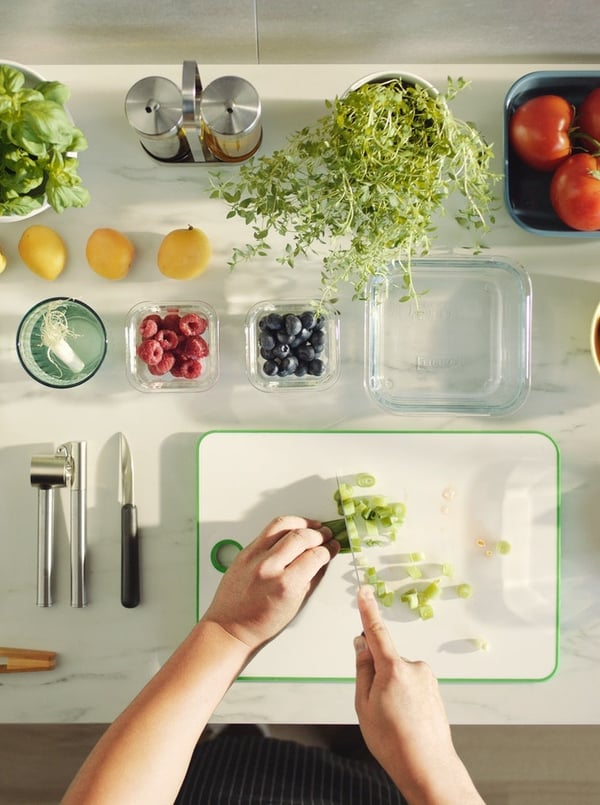 Zwei Hände schneiden Gemüse auf einer Arbeitsfläche, auf der Utensilien, Behälter und Gemüse rechtwinklig angeordnet sind.