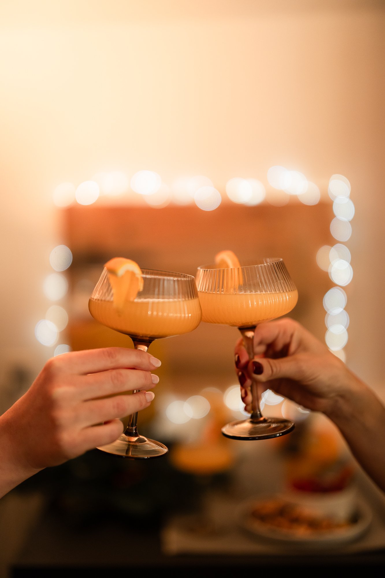 Zwei Hände stossen mit Champagnergläsern an, die mit orangefarbenen Cocktails gefüllt und mit Orangenzesten garniert sind. Warme, festliche Atmosphäre mit sanftem, schimmerndem Licht im Hintergrund.