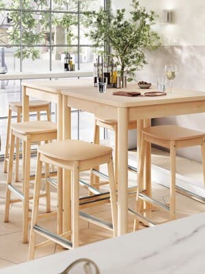 Zwei hohe Tische und fünf Hochstühle aus hellem Holz in einem lichtdurchfluteten Café. 