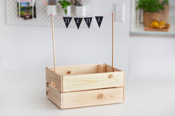 Zwei Holzstäber sind an einer KNAGGLIG Kiste befestigt. Dazwischen hängt eine schwarze Wimpelkette mit der Aufschrift MAMA. 