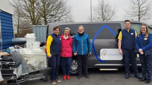 Zwei IKEA-Mitarbeitende in blauer und gelber Kleidung stehen mit zwei Vertreter:innen des Vereins Lichtzeichen e.V. vor einem grauen Transporter mit Vereinslogo. Um sie herum befinden sich Einkaufswagen mit Haushaltswaren und Kartons.