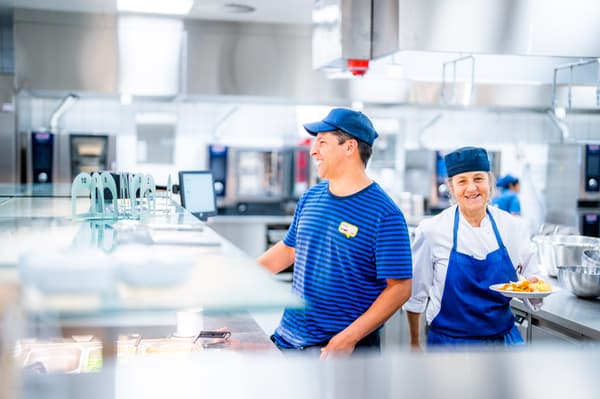 Zwei IKEA Mitarbeitende in dunkelblauen Uniformen lachen, während sie eine kleine Essenspause machen.