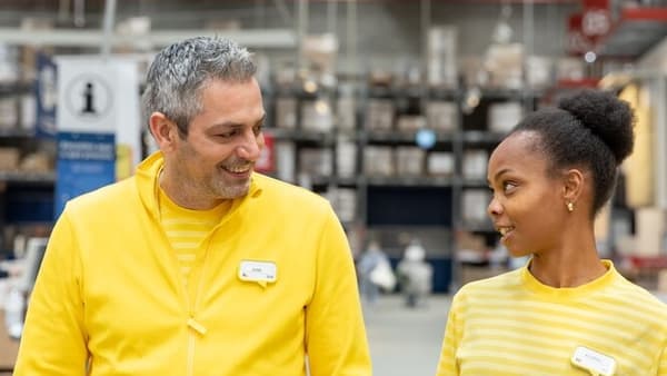 Zwei IKEA-Mitarbeitende in gelber Uniform unterhalten sich freundlich in einem Lagerbereich des IKEA-Einrichtungshauses.