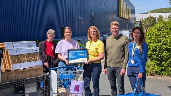 Zwei IKEA Mitarbeitende und drei Vertreter*innen des DRK-Projekts HENRI stehen gemeinsam vor dem IKEA Gebäude; in der Mitte wird ein großer IKEA Siegen Spendenscheck über 5.000 Euro präsentiert. Vor ihnen stehen Einkaufswagen voller Sachspenden wie Stühle, Boxen und weitere Einrichtungsgegenstände.
